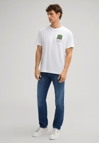 Weißes Baumwoll-T-Shirt mit einem Druck auf der Vorderseite, der grüne Kakteen zeigt. Kombiniert mit blauen Jeans und weißen Turnschuhen. Lässiger Schnitt und kurze Ärmel.