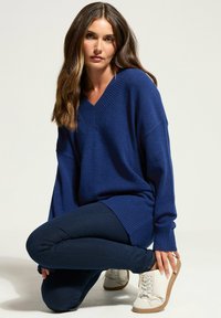 Navyblauer V-Ausschnitt-Pullover mit geripptem Saum und Bündchen, kombiniert mit dunkelblauen Skinny Jeans und weißen Sneakers mit strukturierter Sohle.