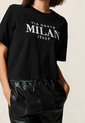 Personne portant un t-shirt court noir avec le texte "Via Dante Milan Italy" et un pantalon en cuir noir à cordon avec les mains dans les poches.