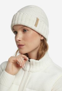 Biała dziana czapka typu beanie o prążkowanej fakturze z skórzaną naszywką. Model nosi białą kurtkę z wysokim kołnierzem, częściowo rozpiętą.