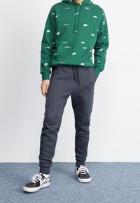 Vihreä huppari, jossa on valkoisia Nike-logoja, yhdistettynä tummanharmaisiin collegehousuihin. Musta-valkoiset ruutukengät. Yksinkertainen, rento urheiluvaatetus.