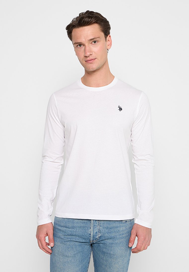 T-shirt bianco a maniche lunghe in cotone, con scollatura rotonda e un piccolo logo nero sul lato sinistro del petto. Abbinato a jeans blu.