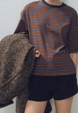Person iført brun og blåstribet t-shirt og sorte shorts, der holder en stor brun tekstureret frakke over den ene arm mod en neutral baggrund.