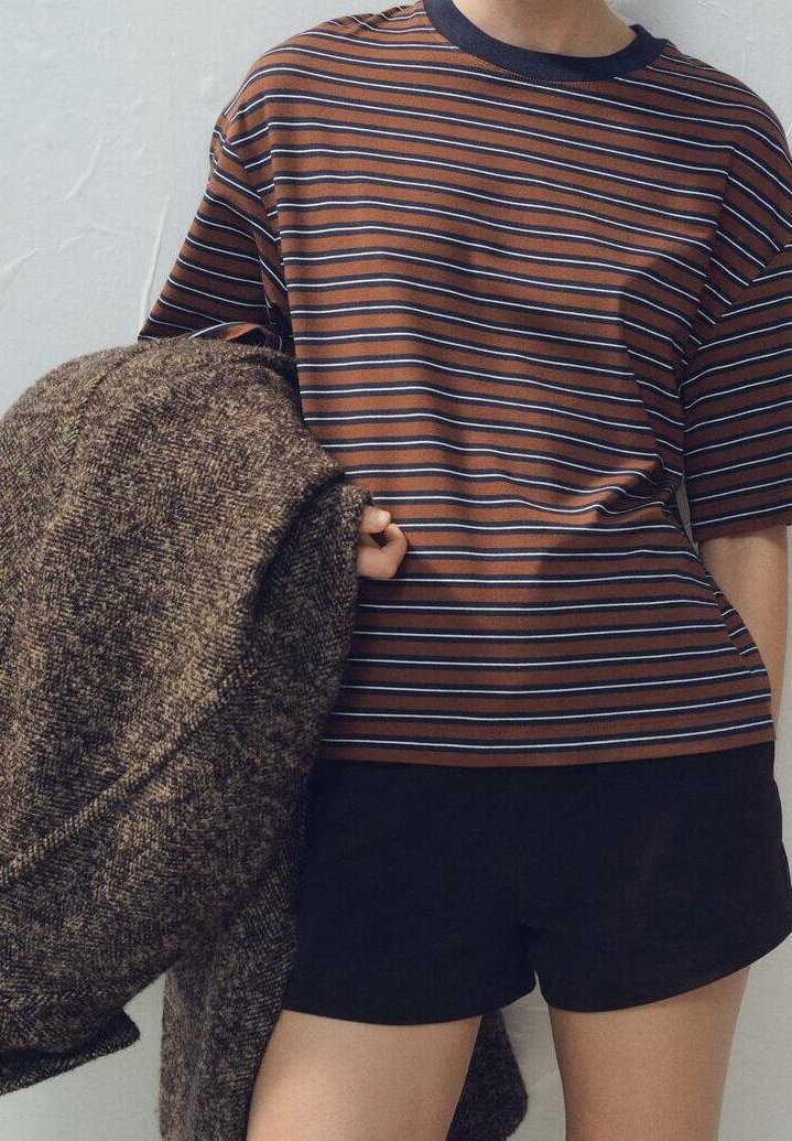 Personne portant un t-shirt rayé marron et bleu et un short noir, tenant un grand manteau marron texturé sur un bras, sur fond neutre.