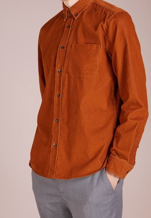 Mann trägt ein rostorangefarbenes Cordhemd mit Brusttasche und graue Hose, Hand in der Tasche, vor schlichtem Hintergrund.