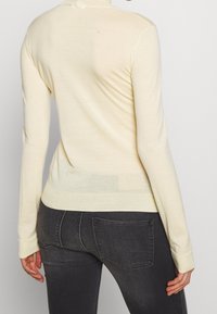 Femme portant un pull à manches longues crème et un jean gris foncé ajusté, vue de dos sur un fond uni.