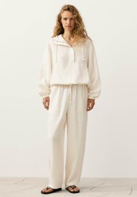 Femme aux cheveux bouclés portant un sweat à capuche ample couleur crème et un pantalon large assorti, debout sur un sol clair devant un fond uni.