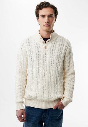 Young man with curly hair wearing a cream cable-knit sweater with wooden buttons and blue jeans standing against a white background.