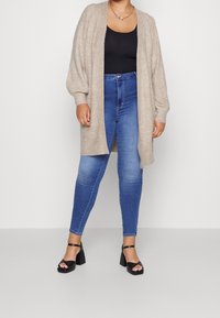 Cardigan lungo beige in materiale morbido, con struttura a coste, design frontale aperto e maniche larghe, abbinato a jeans blu a vita alta e tacchi neri con cinturini.