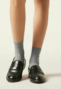 Mocasines de cuero negro con un acabado suave, combinados con calcetines grises de canalé. Los zapatos presentan un detalle de doble hebilla en la parte superior.