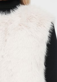 Gilet en fausse fourrure de couleur rose clair doux avec une texture duveteuse. Les bords sont légèrement ébouriffés. Porté par-dessus un pull à col roulé noir.