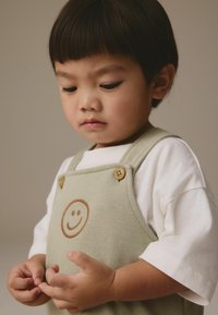 Salopette en coton verte avec une broderie de smiley, boutons en bois, portée sur un T-shirt blanc. Les mains de l'enfant sont réunies.