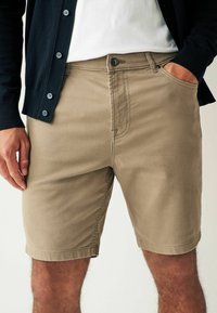 Shorts en coton beige à coupe droite, avec poches avant et fermeture à bouton. Associés à un cardigan bleu marine et un t-shirt blanc.
