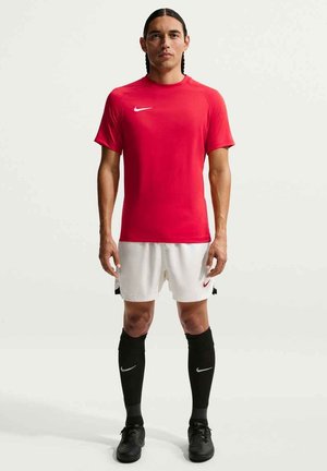 Atleta con capelli intrecciati in piedi, con maglia rossa a maniche corte, pantaloncini bianchi, calze nere fino al ginocchio e tacchetti neri su sfondo semplice.