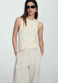 Débardeur en tricot crème sans manches associé à un pantalon large beige et blanc à rayures, avec des plis, des poches latérales et une coupe décontractée.