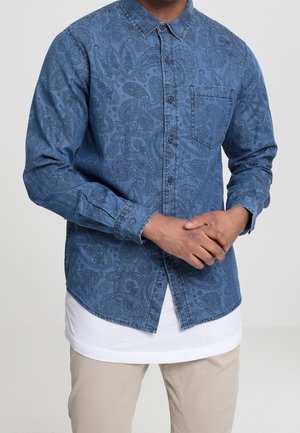 Mężczyzna nosi niebieską koszulę z długim rękawem z wzorem paisley, zapinaną na guziki, na białym t-shircie i beżowych spodniach, z rękami złożonymi przed sobą.