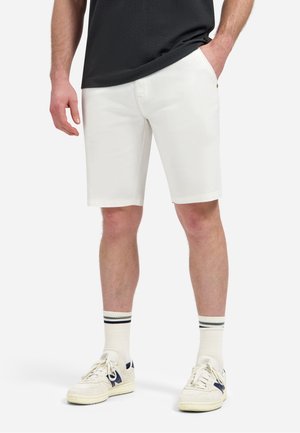 Man draagt witte knielange shorts, witte sneakers met donkerblauwe accenten, witte sokken met zwarte strepen en een zwart shirt.