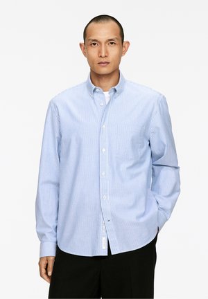 Chemise à boutons rayée bleu et blanche en tissu à texture lisse, dotée d'un col, d'une poche avant et d'un ourlet arrondi.