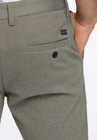 Olivgrüne maßgeschneiderte Hosen aus strukturiertem Stoff, mit einer Gesäßtasche mit einem schwarzen Knopf und einem kleinen Markenschild am Bund.