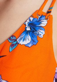 Oranžová látka se modrými květy hibiskusu a zelenými listy. Obsahuje tenké ramínka s dekorativními prvky. Hladký povrch.