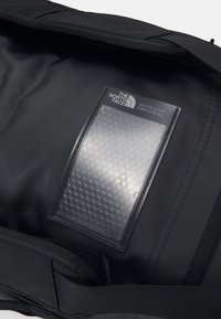 Sac de voyage noir avec une surface lisse et texturée et une poche transparente présentant un motif hexagonal. Logo de la marque visible à l'intérieur.