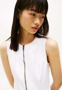 Biała sukienka bez rękawów z wysokim kołnierzem, zamkiem błyskawicznym z przodu, kontrastującymi przeszyciami i miękką fakturą. Zbliżenie na górną część ciała.