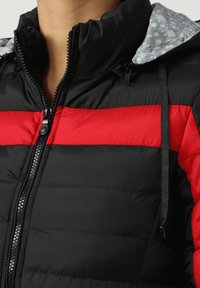 Chaqueta acolchada negra con rayas horizontales rojas, cierre de cremallera, capucha ajustable con forro gris con diseño y detalles de costuras visibles.