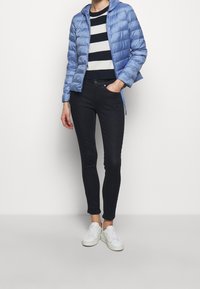 Jasnoniebieska kurtka puchowa z kapturem, czarno-biała sweter w paski, ciemne dżinsy o wąskim kroju i białe sneakersy. Gładkie tekstury i dopasowany fason.