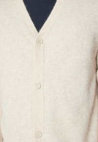 Ljusbeige cardigan i mjukt, texturerat tyg, med V-ringning, framsidans knappar och ribbade detaljer längs kanterna.