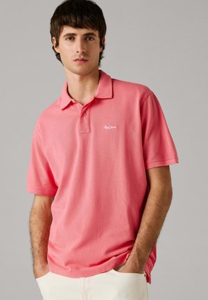 Young man with dark hair wearing a coral pink polo shirt with "Pepe Jeans" logo, hands in pockets, standing against a plain gray background.