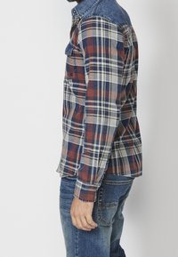 Homme portant une chemise à carreaux bleue, rouge et blanche avec un col en denim et un jean bleu, debout de profil sur fond blanc.