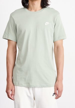 Mężczyzna w jasnozielonej koszulce Nike i białych szortach Nike, stojący na jednolitym białym tle.