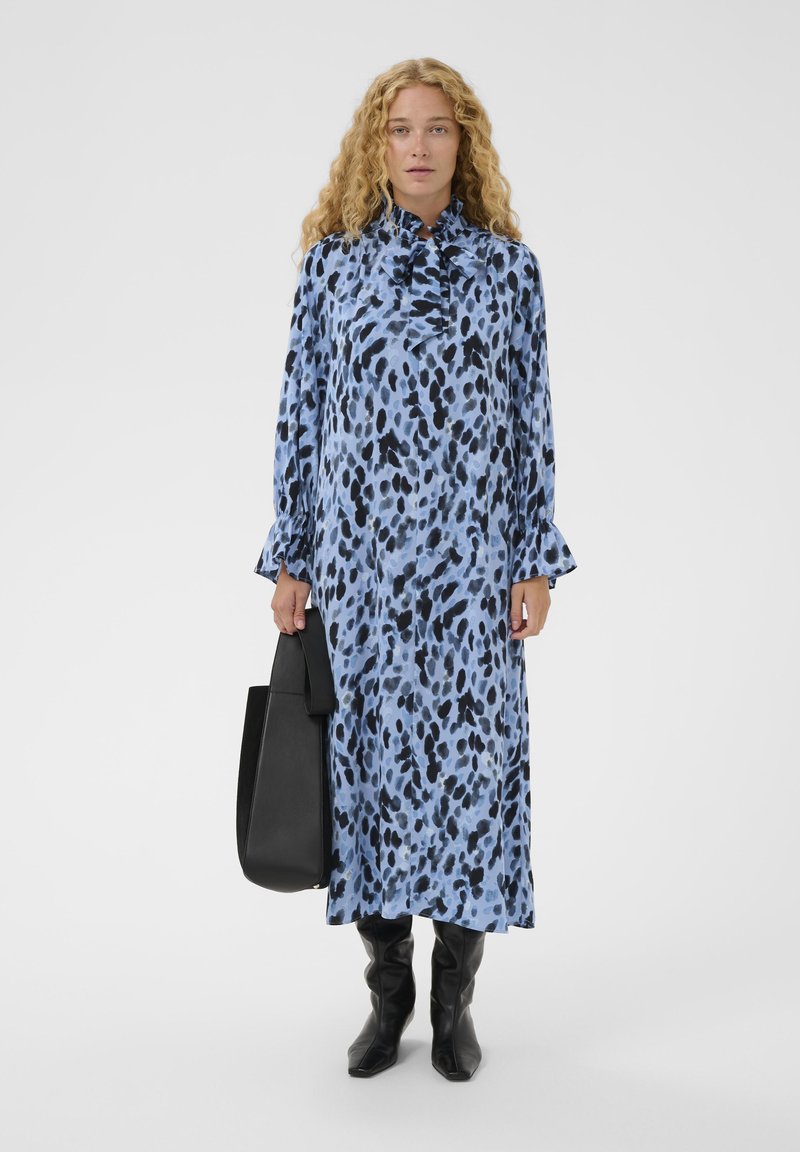 Femeie cu păr creț, îmbrăcată într-o rochie lungă albastre cu pete negre, ținând o geantă mare neagră și cizme negre, stând pe un fundal alb simplu.