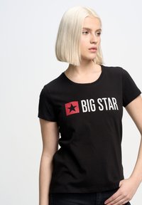 Juoda trumpomis rankovėmis marškinėliai, vaizduojantys raudoną kvadratinį logotipą su juoda žvaigžde ir balta tekstu "BIG STAR" ant priekio. Minkšta medvilnė.