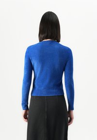 Vrouw met steil donker haar draagt een aansluitende felblauwe langemouwen top en een hooggetailleerde zwarte rok, staande tegen een witte achtergrond.