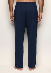 Man som står barfota, sett bakifrån, iklädd löst sittande navyblå byxor mot en enfärgad beige bakgrund.