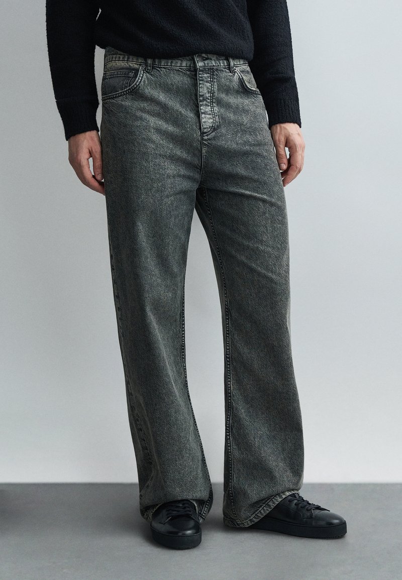 Személy kopott fekete, bő fazonú farmernadrágot, fekete tornacipőt és fekete pulóvert visel, szürke padlón áll, világosszürke fal előtt.