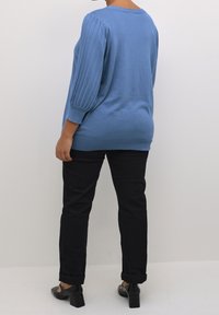 Personne debout, portant un pull à manches côtelées bleu, un pantalon noir et des chaussures à talons noires, tournée de dos sur un fond blanc uni.