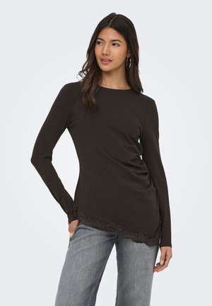 Kvinde med langt mørkt hår iført sort langærmet top med blondekant og lysegrå jeans, kigger sidelæns med hånden på hoften.