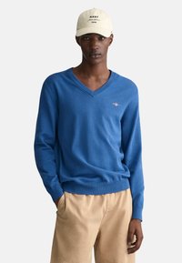 Blauer V-Ausschnitt-Pullover aus weichem Strickmaterial mit gerippten Bündchen und Saum, kombiniert mit khakifarbenen Shorts. Mütze mit aufgedrucktem Logo der Marke.