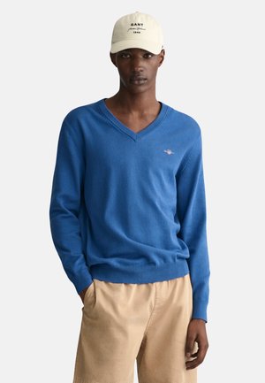 Blue V-neck sweater made of soft knit material, featuring ribbed cuffs and hem, paired with khaki shorts. Cap with brand logo displayed.