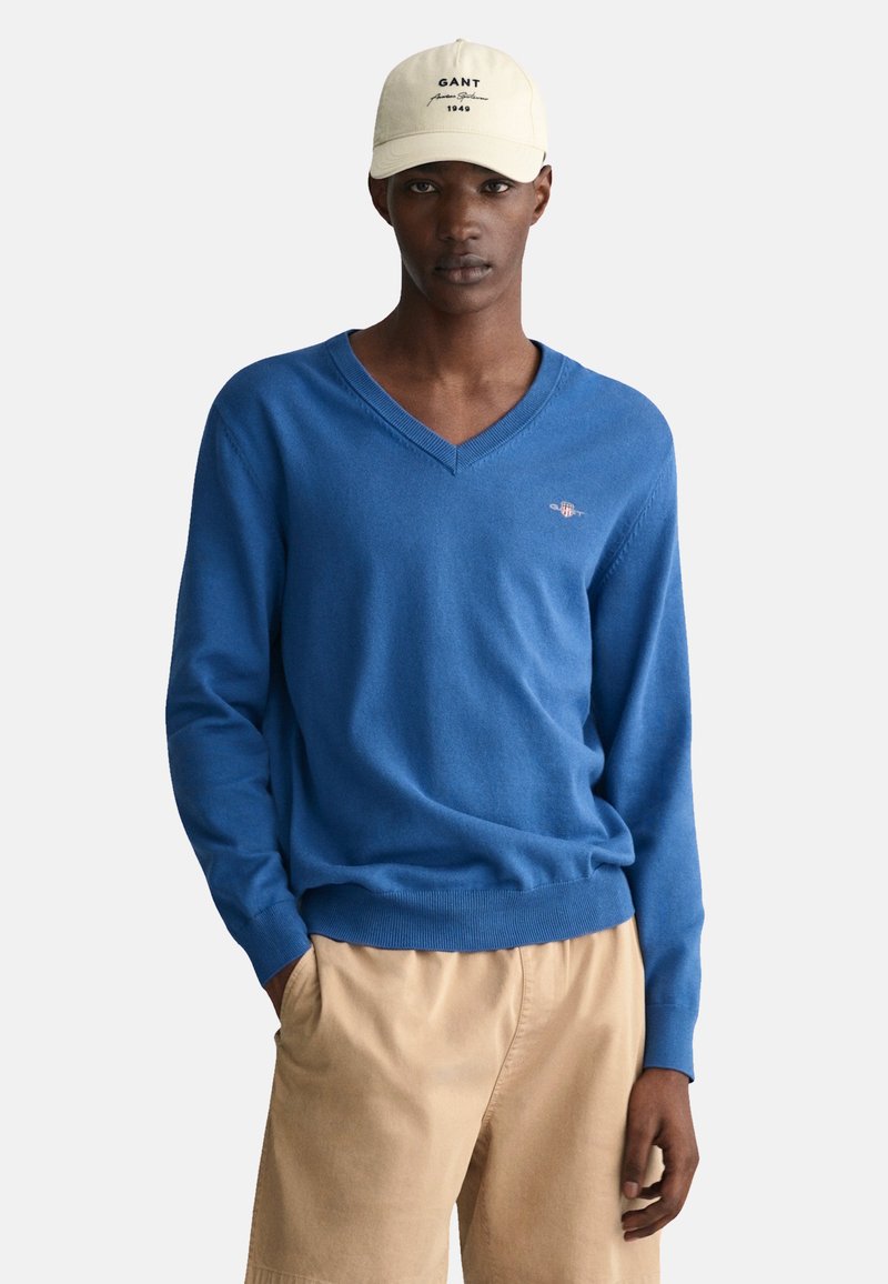 Blauer V-Ausschnitt-Pullover aus weichem Strickmaterial mit gerippten Bündchen und Saum, kombiniert mit khakifarbenen Shorts. Mütze mit aufgedrucktem Logo der Marke.