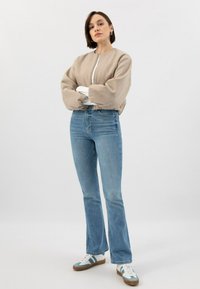 Giubbotto bomber beige accorciato con maniche a sbuffo e zip frontale, abbinato a jeans svasati azzurri chiari e sneakers sportivi grigio-blu.