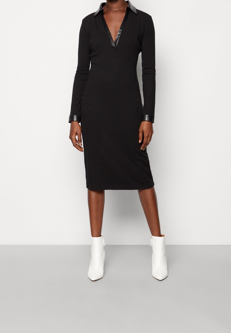 Femme portant une robe midi noire à manches longues avec col et poignets en cuir, associée à des bottines blanches, debout devant un fond uni.