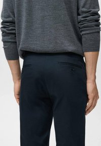 Mörkblå byxor med två bakfickor och knappdetaljer, i kombination med en grå texturerad tröja med uppvikta ärmar.