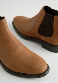 Botas de tobillo de cuero marrón con un acabado suave, paneles laterales elásticos y una suela de goma plana. Presentan una puntera redondeada y detalles cosidos.