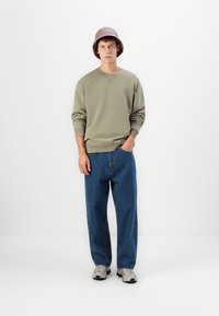 Joven con sudadera verde, jeans anchos azules, zapatillas grises y un sombrero de cubo malva de pie contra un fondo blanco.