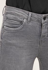 Jeans grises con un corte ajustado, que presentan un bolsillo frontal, un lazo para un cinturón y un cierre de botón. La tela tiene un acabado liso, ligeramente texturizado.