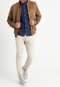 Man draagt een bruine suède jas, een navy blouse met knopen, beige broek en grijze sneakers, staand tegen een witte achtergrond.