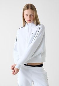 Weißes Sweatshirt mit Reißverschluss, schwarzen Paspel-Details, lockerem Schnitt und gerippten Bündchen. Kombiniert mit weißen Hosen mit schwarzem Logo-Bund.